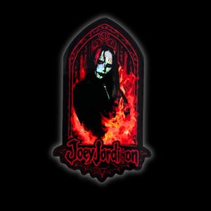 May include: A black patch with a portrait of Joey Jordison, his face painted, surrounded by red flames. The patch is in a gothic arch shape and has the name "Joey Jordison" written in red script.