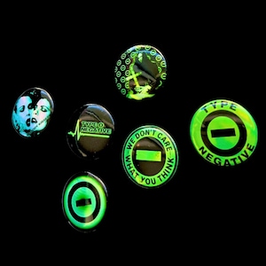 Peut inclure: Un ensemble de six badges ronds avec des visuels du groupe Type O Negative. Les motifs présentent des graphismes vert néon et noir, avec les inscriptions "Type O Negative", "Bloody Kisses" et "We Don't Care What You Think", ainsi qu'un symbole moins.