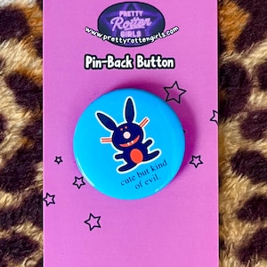 May include: A blue pin-back button with a cartoon bunny design and the text "cute but kind of evil." The button is on a purple card with the text "Pin-Back Button" and the website "prettyrottengirls.com". The card has star graphics.