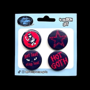 May include: A set of four button pins on a light blue card. The pins are dark blue with red graphics and text, including a star, a girl, and the phrases "get lost stay lost" and "HOT GOTH". The card has the text "4 BUTTON SET".