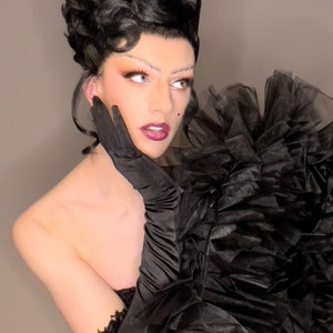 May include: A person with dark hair styled in a bouffant, wearing black lace gloves and a black tulle ruffle top. The person has dramatic makeup with dark eyeliner and red lipstick.