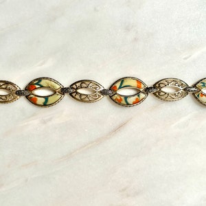 May include: A gold bracelet with a delicate filigree design and oval-shaped links. Each link features a colorful floral pattern on a cream background.