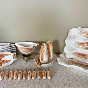 May include: A ceramic dinnerware set featuring a corn-on-the-cob motif, including several long rectangular plates, a gravy boat on a metal stand, salt and pepper shakers, and a collection of small corn-handled metal skewers for serving corn.