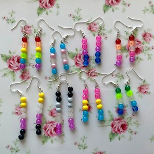 May include: A set of 10 pairs of beaded earrings, each pair featuring a different color combination. The earrings are made with small, round beads in various colors, including yellow, red, blue, green, white, black, and purple. The earrings are strung on silver-toned earring hooks.