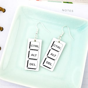 May include: A pair of white rectangular earrings with the words CTRL, ALT, and DEL printed on them in black. The earrings are hanging on silver hooks. The earrings are on a light green surface.