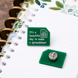 May include: Green enamel pin with the text "It's a beautiful day to make a spreadsheet" and an XLS icon. The pin is on a white notebook with a floral design.