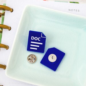 May include: A square, light blue ceramic dish holds a blue DOC file pin, a blue envelope pin, and a silver pin. The DOC file pin has the word "DOC" and three lines. The pins are on a white notebook with gold spiral binding.