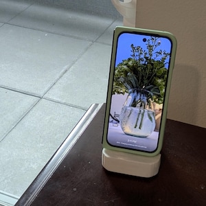 May include: A light green phone case with a white phone stand. The phone screen shows a photo of a vase with green flowers.