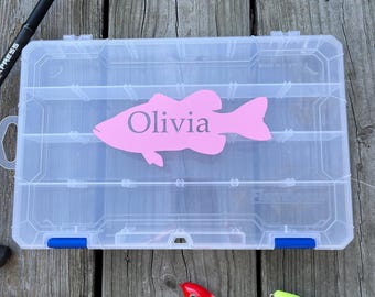 May include: A clear plastic fishing tackle box with a pink fish-shaped decal that reads "Olivia." The box has blue latches and is surrounded by fishing lures and a fishing rod. The lures are red, white, and black.