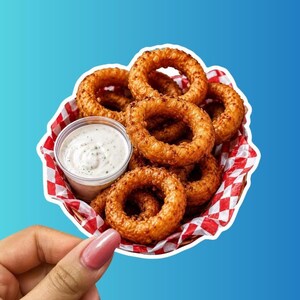 May include: A basket of golden-brown, crispy onion rings served with a small plastic container of creamy white dipping sauce. The basket is lined with red and white checkered paper, and the entire arrangement is presented as a die-cut sticker.