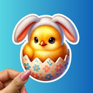 May include: A yellow chick wearing a white bunny ear hat sits inside a pastel pink and blue Easter egg with white flowers.