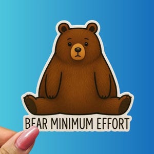May include: A brown bear sticker with a white border, sitting with its legs out. The bear has a light brown snout and a black nose. The text "BEAR MINIMUM EFFORT" is printed in black on a white banner below the bear. The background is a gradient of blue.