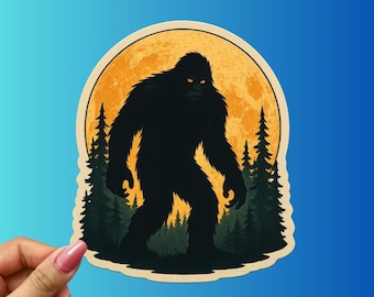 May include: A Bigfoot sticker featuring a black silhouette of the creature against a full orange moon. The design includes dark green pine trees and a beige border, set against a gradient blue background.