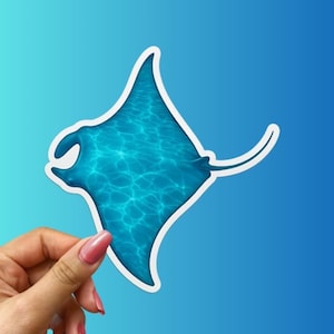May include: A sticker featuring a stylized manta ray with a textured, water-like pattern in shades of blue. The manta ray is outlined in white against a gradient blue background. The sticker is held by a hand.