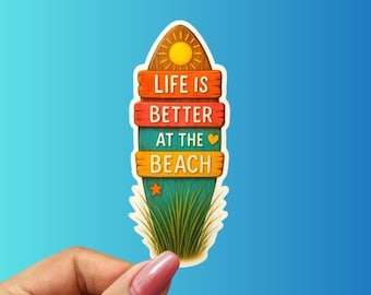 Puede incluir: Una colorida pegatina con el texto "LIFE IS BETTER AT THE BEACH". El diseño presenta un sol, una estrella y hierba, con las palabras en tablones de madera. La pegatina está sujeta por una mano sobre un fondo azul.
