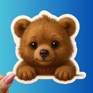 May include: A brown teddy bear sticker with a smiling expression and large, dark eyes. The bear's fur is detailed, and it appears to be peeking over a white border against a blue background. The sticker is held by a hand with a pink nail.