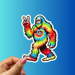 May include: A tie-dye Bigfoot sticker with a peace sign on its chest. The sticker is white with a colorful tie-dye pattern in shades of blue, green, yellow, orange, and red.