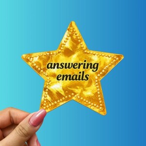 May include: A gold star-shaped sticker with the words "answering emails" in black text. The star has a textured surface and is held by a hand with a pink nail. The background is a gradient of blue.