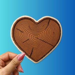 May include: A heart-shaped wooden coaster with a tree ring design. The coaster features a brown interior with concentric circles and cracks, surrounded by a white border. The background is a gradient of blue.