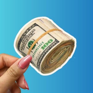 May include: A thick roll of United States one hundred dollar bills secured by a single tan rubber band. The currency features the standard green and off-white paper texture with visible serial numbers and the prominent number 100 printed on the bills.