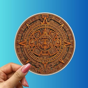 May include: A round, detailed sticker featuring the Aztec calendar stone design. The sticker is primarily brown and gold, with intricate carvings and a central face. The background is a gradient of blue.