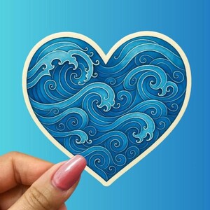 May include: A heart-shaped sticker featuring a stylized ocean wave design in shades of blue. The waves have intricate swirling patterns and are outlined in a darker blue. The sticker has a cream-colored border.