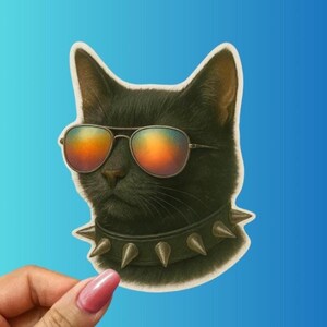 May include: A sticker featuring a black cat wearing aviator sunglasses with orange and yellow lenses and a studded collar. The cat's fur is dark, and the sunglasses have a silver frame. The sticker has a white border and is held by a hand.