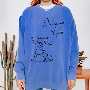Pu&ograve; includere: Una felpa blu con una grafica di una donna che cammina con una valigia e il testo "Airplane Mode" in nero.