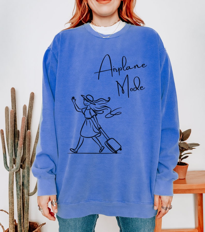 Pu&ograve; includere: Una felpa blu con una grafica di una donna che cammina con una valigia e il testo "Airplane Mode" in nero.