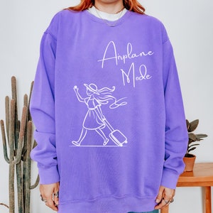 Puede incluir: Sudadera morada con un gr&aacute;fico blanco de una mujer con sombrero tirando de una maleta con el texto "Airplane Mode" en la parte delantera.