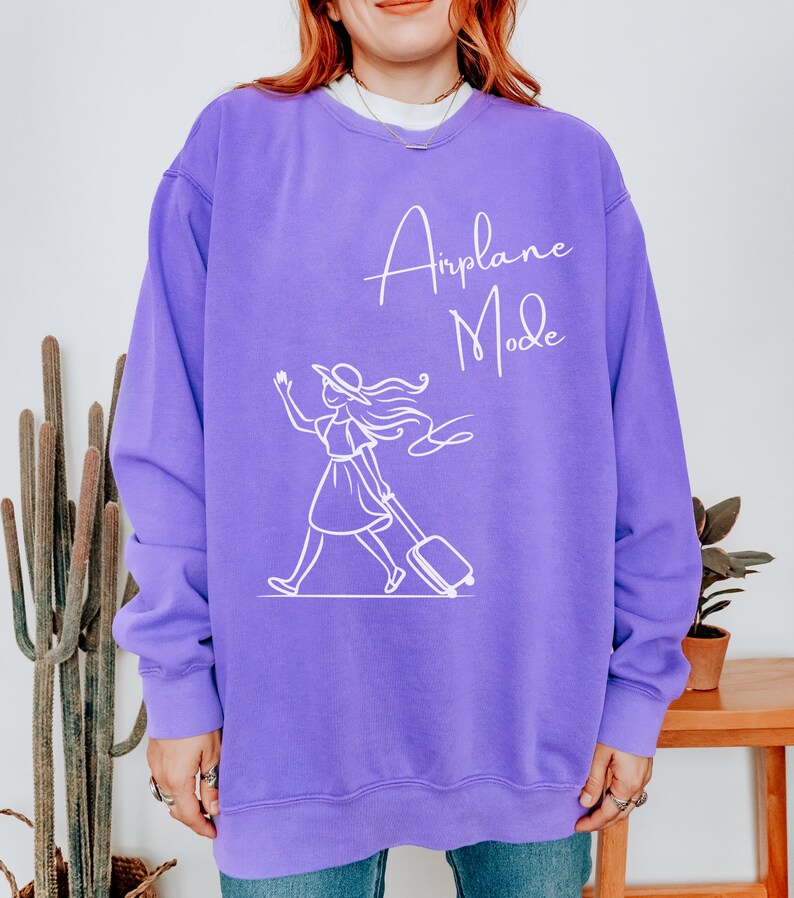 Puede incluir: Sudadera morada con un gr&aacute;fico blanco de una mujer con sombrero tirando de una maleta con el texto "Airplane Mode" en la parte delantera.