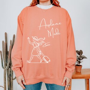 Puede incluir: Una sudadera de color coral con el texto "Airplane Mode" en letras blancas. Debajo del texto hay un dibujo lineal blanco de una mujer con una maleta.