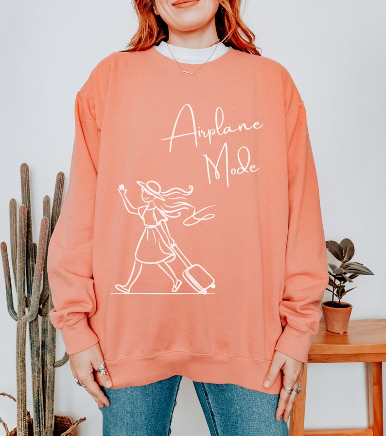 Puede incluir: Una sudadera de color coral con el texto "Airplane Mode" en letras blancas. Debajo del texto hay un dibujo lineal blanco de una mujer con una maleta.