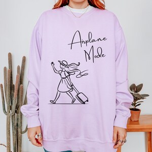 Pu&ograve; includere: Una felpa color lavanda con una grafica nera di una donna con un cappello che tira una valigia con il testo "Airplane Mode" su di essa.