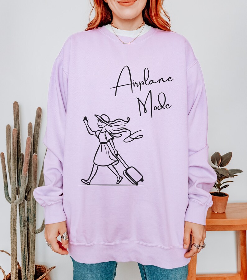 Pu&ograve; includere: Una felpa color lavanda con una grafica nera di una donna con un cappello che tira una valigia con il testo "Airplane Mode" su di essa.