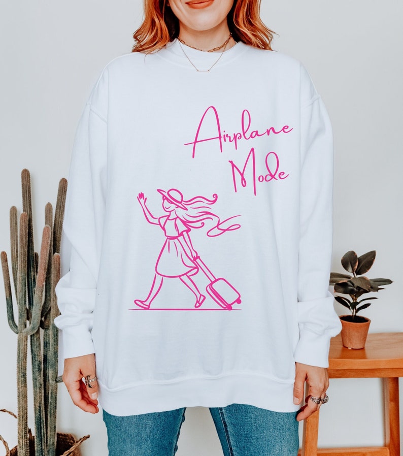May include: White sweatshirt with a pink graphic of a woman in a hat pulling a suitcase with the text "Airplane Mode" in pink cursive.