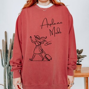 Pu&ograve; includere: Una felpa color granata con una grafica nera di una donna con un cappello che tira una valigia con la scritta "Airplane Mode" stampata su di essa.