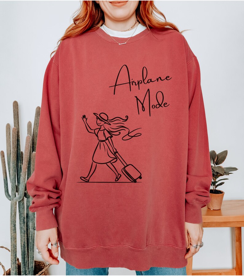 Pu&ograve; includere: Una felpa color granata con una grafica nera di una donna con un cappello che tira una valigia con la scritta "Airplane Mode" stampata su di essa.