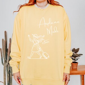 Puede incluir: Sudadera amarilla con un gr&aacute;fico blanco de una mujer con una maleta y el texto "Airplane Mode".