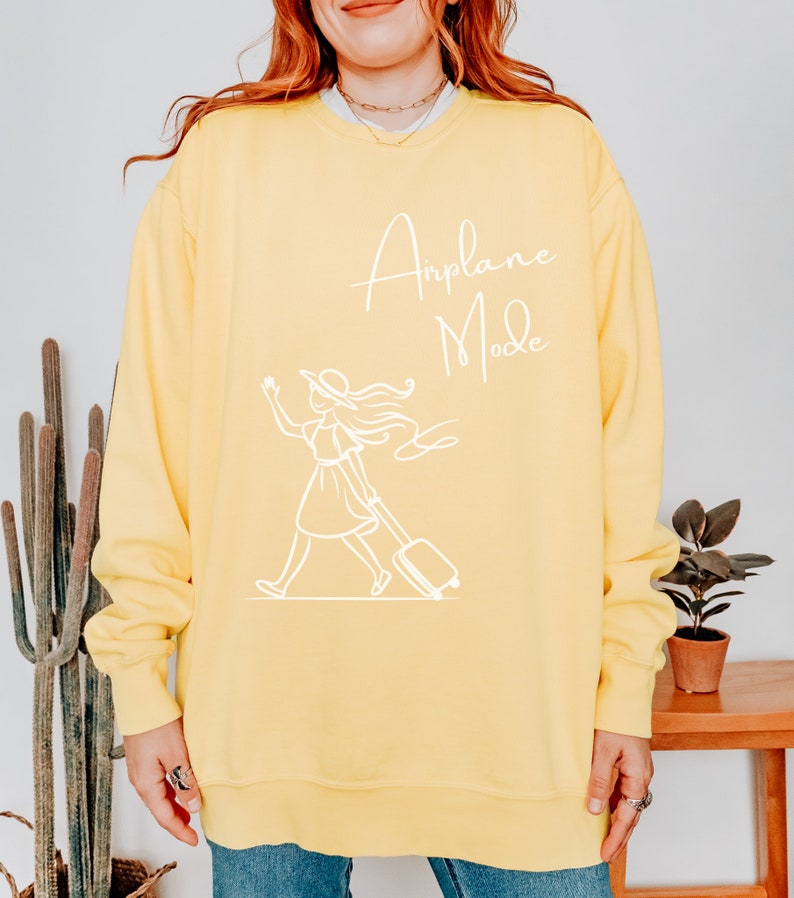 Puede incluir: Sudadera amarilla con un gr&aacute;fico blanco de una mujer con una maleta y el texto "Airplane Mode".