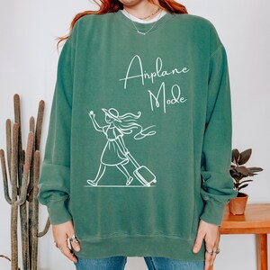 Puede incluir: Una sudadera verde con el texto "Airplane Mode" en letras blancas en cursiva. Debajo del texto hay un dibujo de l&iacute;nea blanca de una mujer con una maleta.