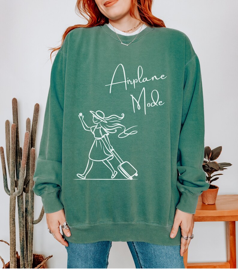 Puede incluir: Una sudadera verde con el texto "Airplane Mode" en letras blancas en cursiva. Debajo del texto hay un dibujo de l&iacute;nea blanca de una mujer con una maleta.