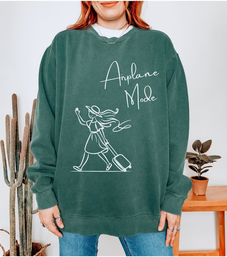 Puede incluir: Una sudadera verde oscuro con el texto "Airplane Mode" en letras blancas cursivas. Debajo del texto hay un dibujo de l&iacute;nea blanca de una mujer con una maleta y un sombrero.
