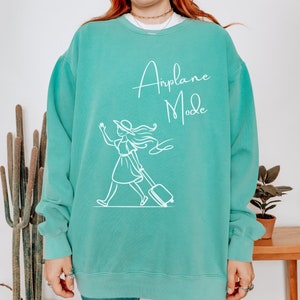 Puede incluir: Una sudadera de color azul turquesa con un gr&aacute;fico blanco de una mujer con un sombrero tirando de una maleta con el texto "Airplane Mode" en la parte delantera.