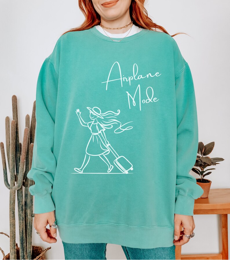 Puede incluir: Una sudadera de color azul turquesa con un gr&aacute;fico blanco de una mujer con un sombrero tirando de una maleta con el texto "Airplane Mode" en la parte delantera.