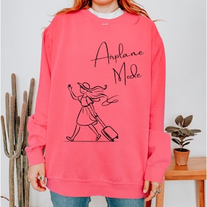 Pu&ograve; includere: Una felpa rosa con una grafica nera di una donna con cappello e vestito che porta una valigia. Il testo "Airplane Mode" &egrave; stampato sulla felpa.