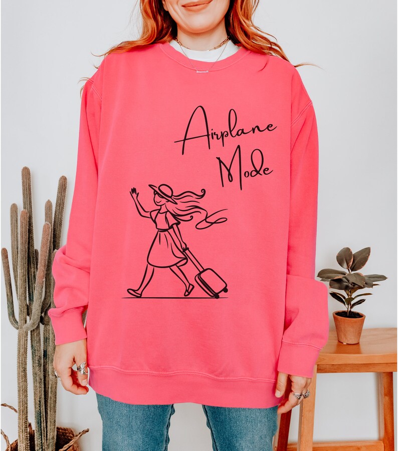 Pu&ograve; includere: Una felpa rosa con una grafica nera di una donna con cappello e vestito che porta una valigia. Il testo "Airplane Mode" &egrave; stampato sulla felpa.