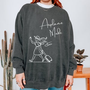 Puede incluir: Una sudadera gris oscuro con un gr&aacute;fico blanco de una mujer con un sombrero tirando de una maleta con el texto "Airplane Mode" encima.