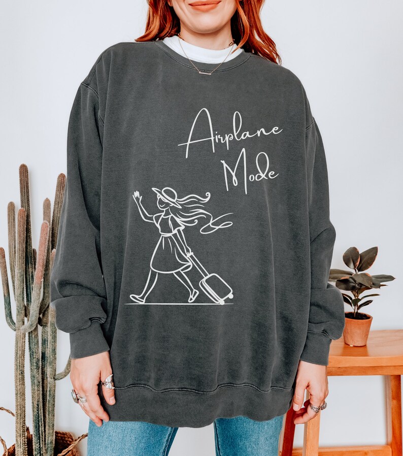Puede incluir: Una sudadera gris oscuro con un gr&aacute;fico blanco de una mujer con un sombrero tirando de una maleta con el texto "Airplane Mode" encima.
