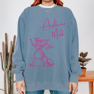May include: A light blue sweatshirt with a pink graphic of a woman pulling a suitcase and the text "Airplane Mode".